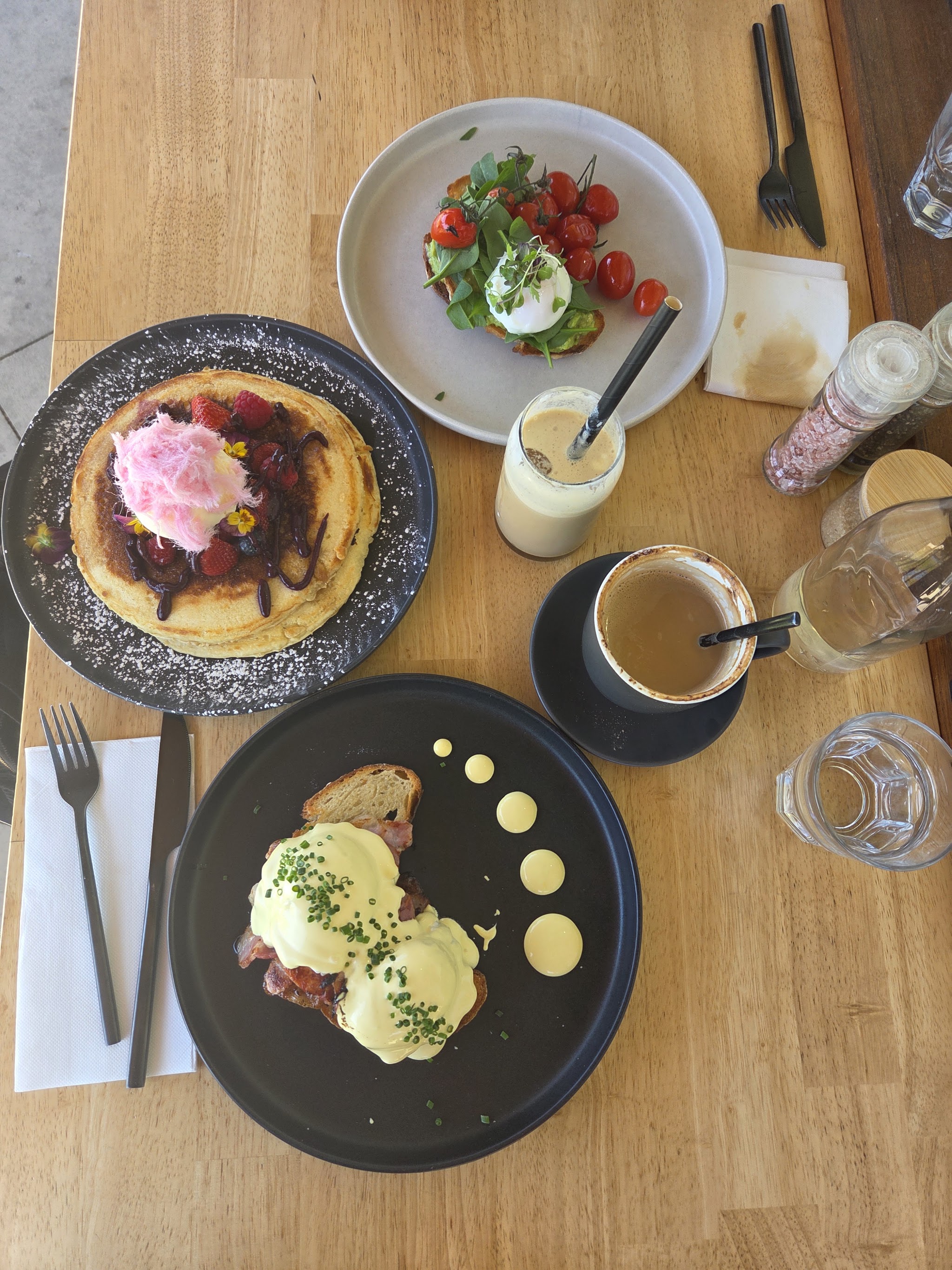 A table spread with pancakes, eggs benedict, smashed avocado, an iced coffee and a flat white